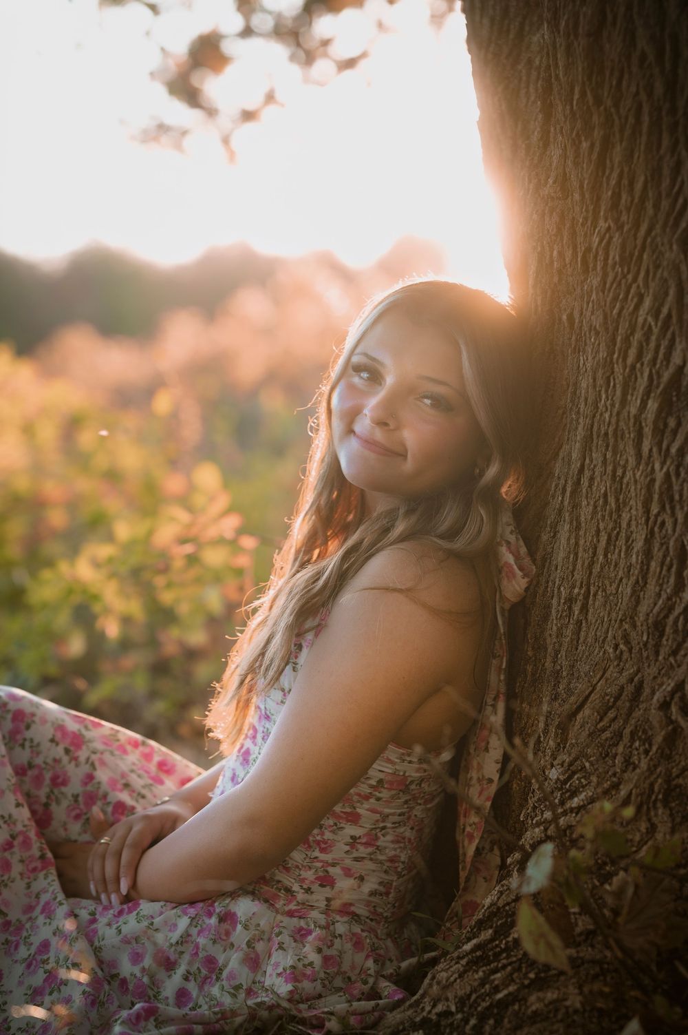A person in a floral dress sits against a tree trunk during golden hour sunset with warm backlit lighting.