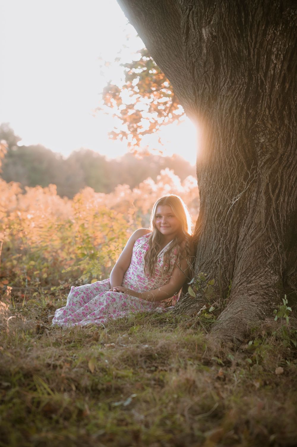 A series of dreamy sunset photos showing someone resting against a large tree trunk in a floral dress.