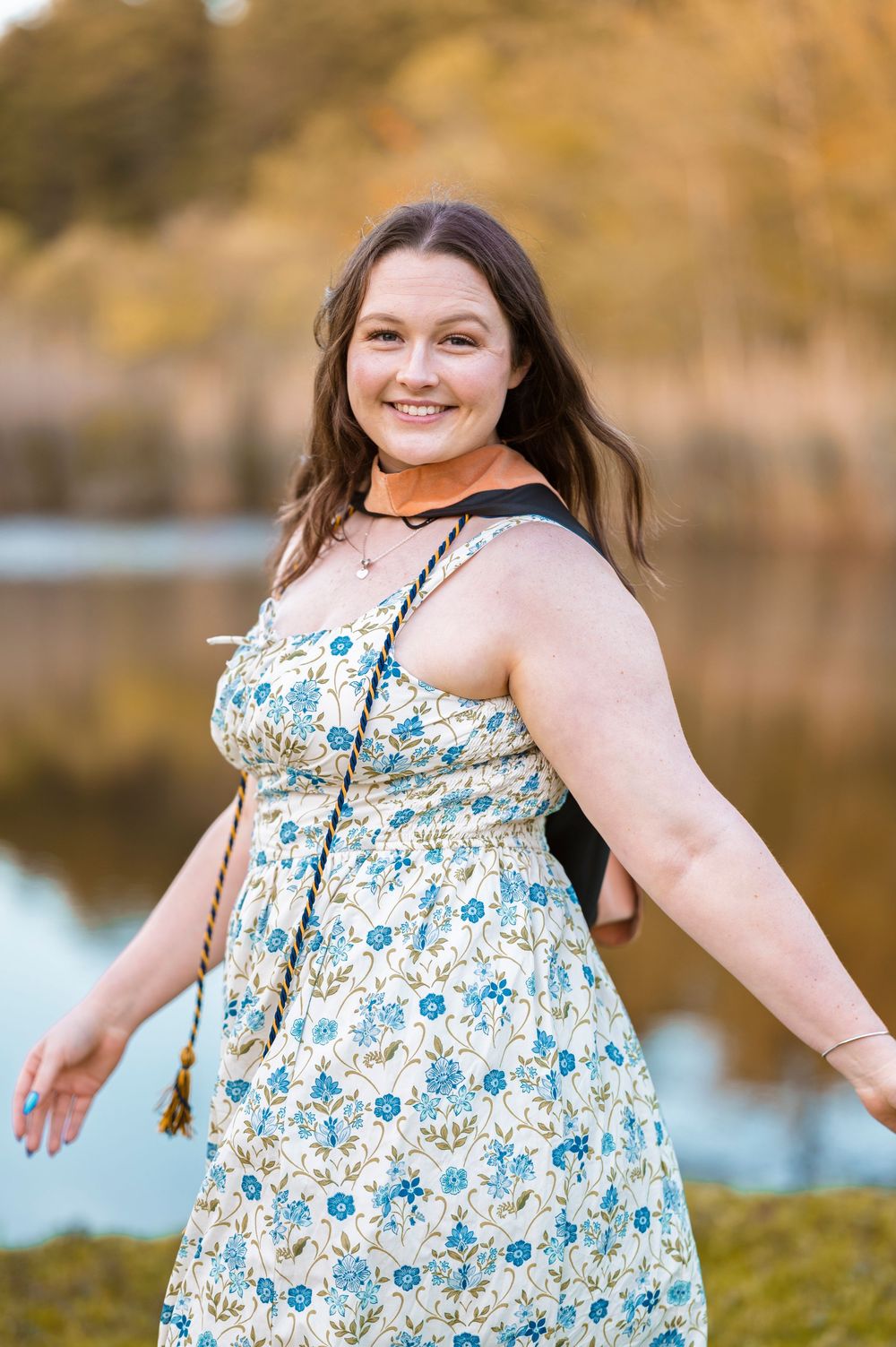 A person wearing a white and blue floral dress poses outdoors near a lake in autumn.
