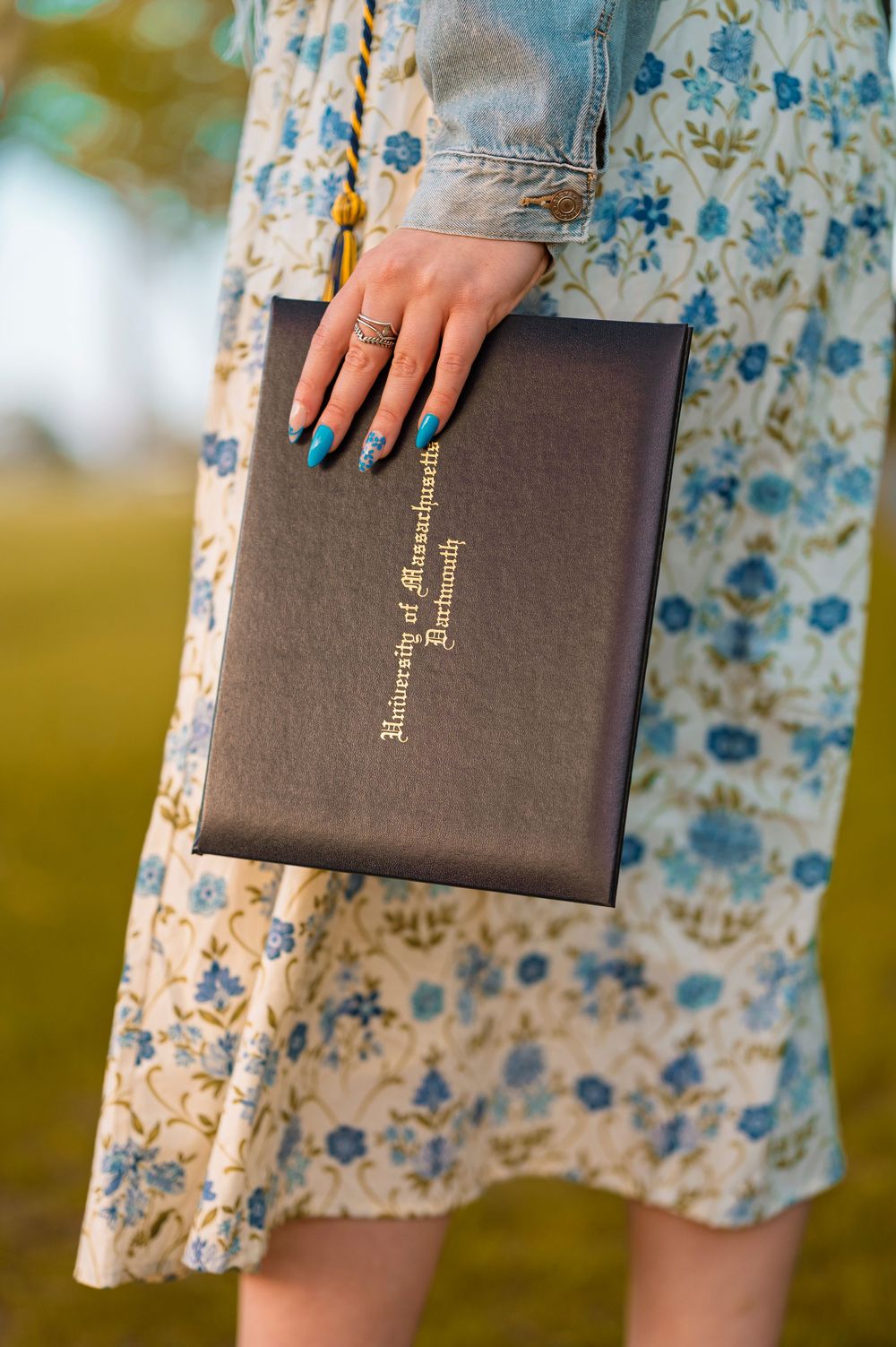 Hand with turquoise nails holds a leather-bound book against a floral print dress outdoors.
