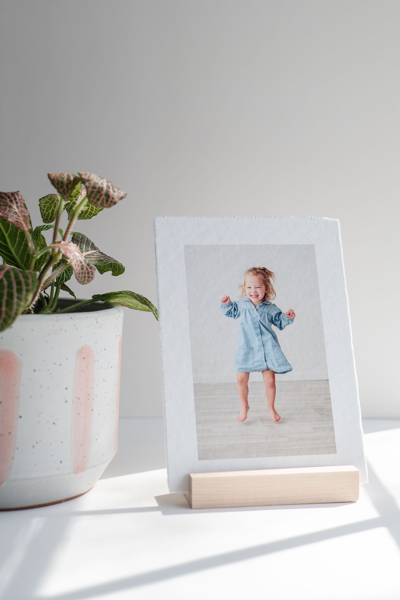 A photo frame on a wooden stand sits next to a potted plant on a white surface with natural lighting.