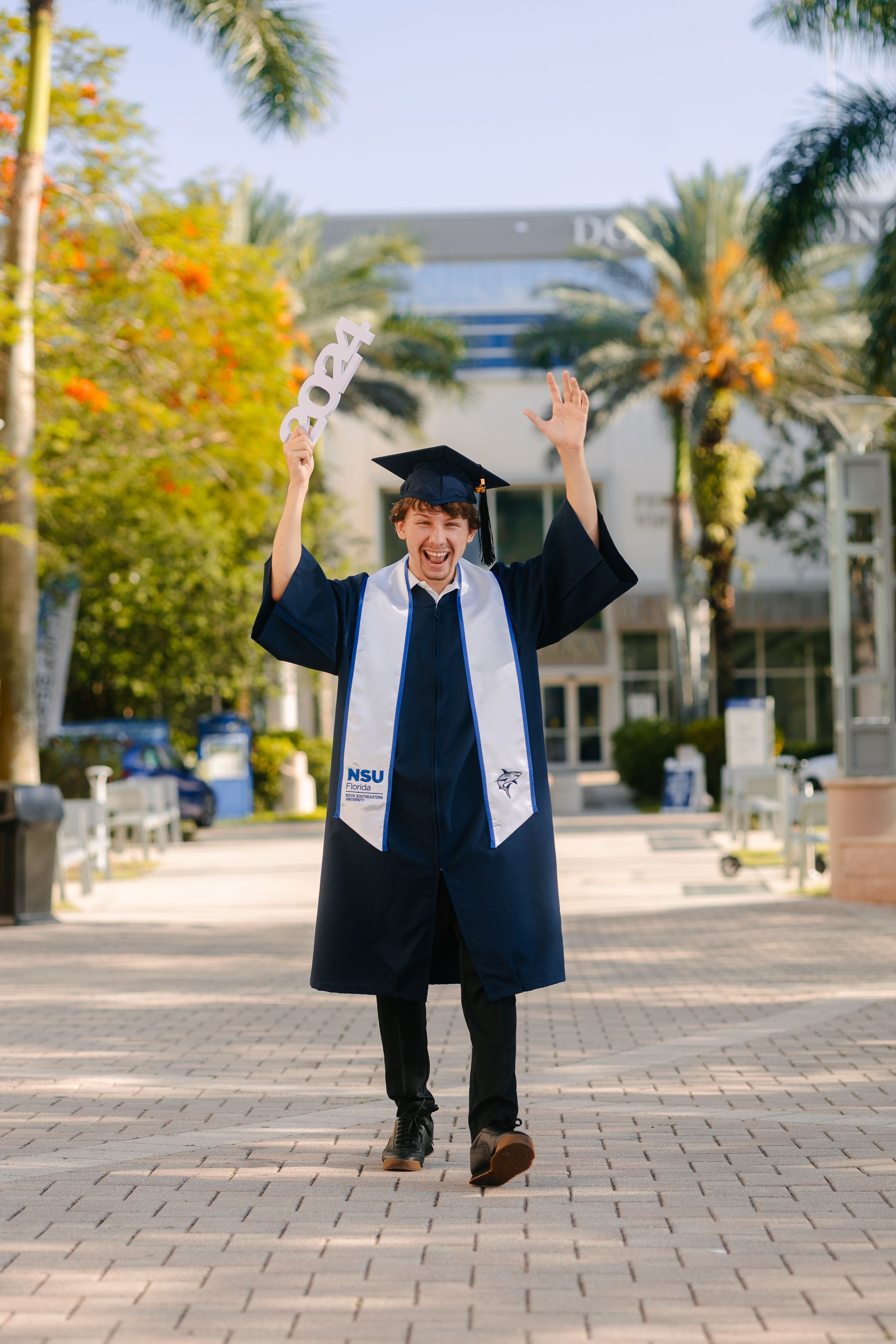 Graduate in gown celebrating outdoors with palm trees, South Florida graduation portrait by Mannyportraits for FIU and UM students.