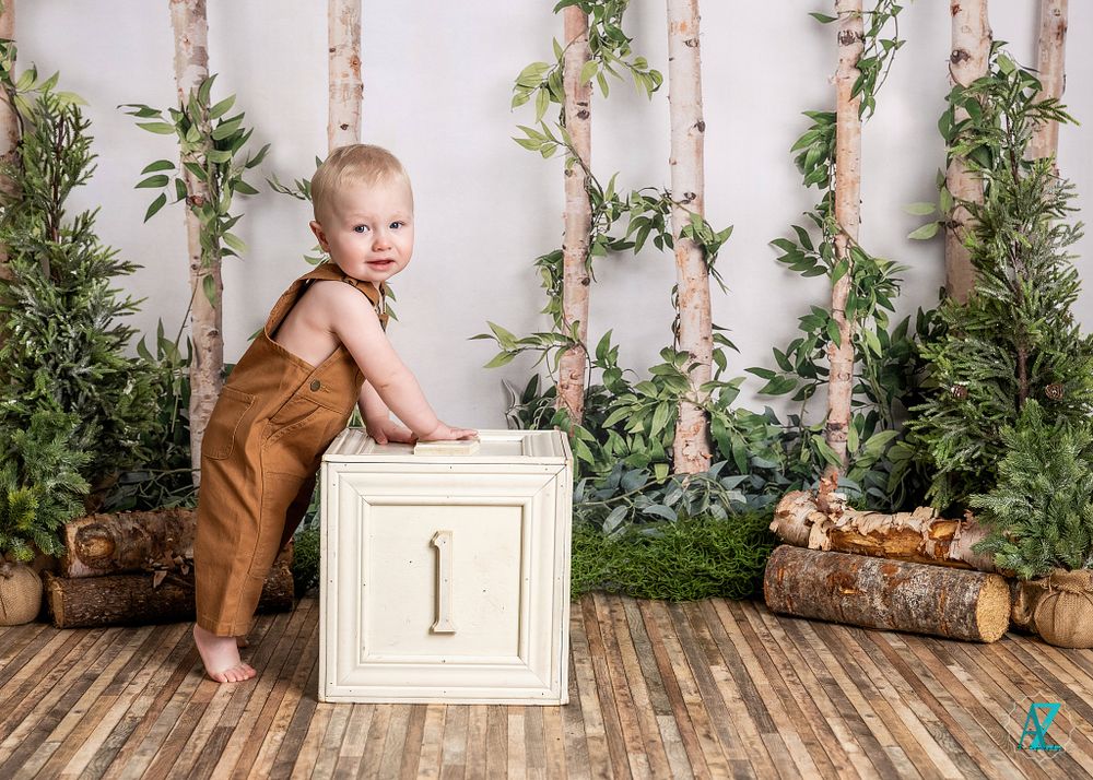Small child in brown overalls stands next to white wooden box in woodland-themed studio setting.