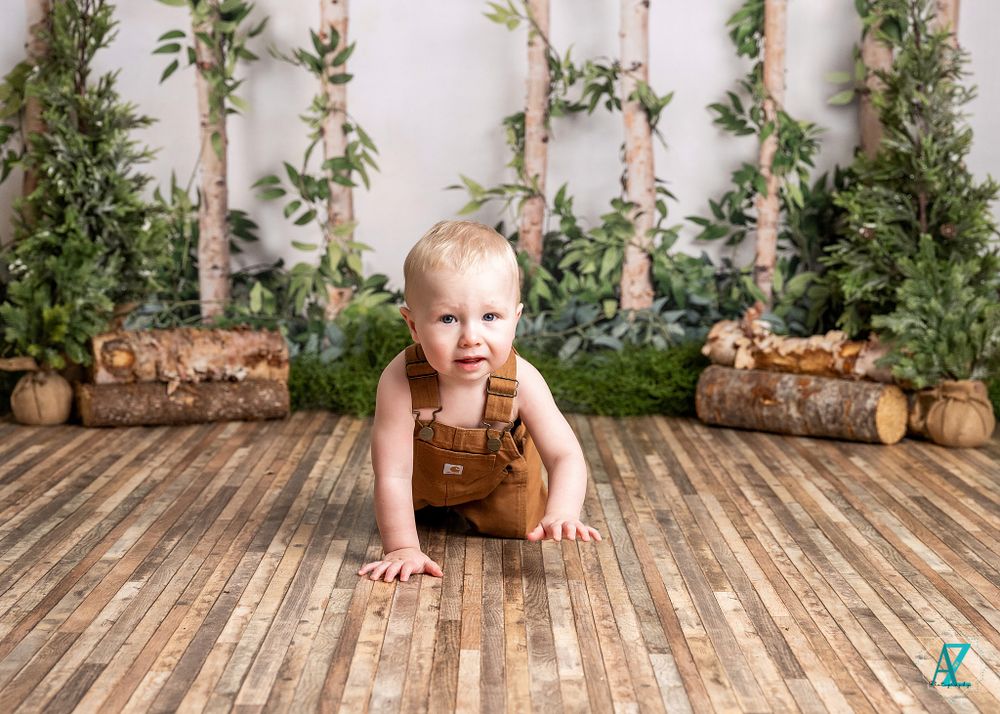 Child in brown overalls crawls on wooden floor with forest-themed backdrop.