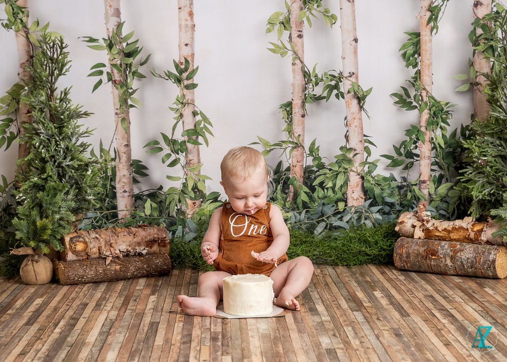 Child in brown overalls sits with white box prop against forest-themed backdrop.