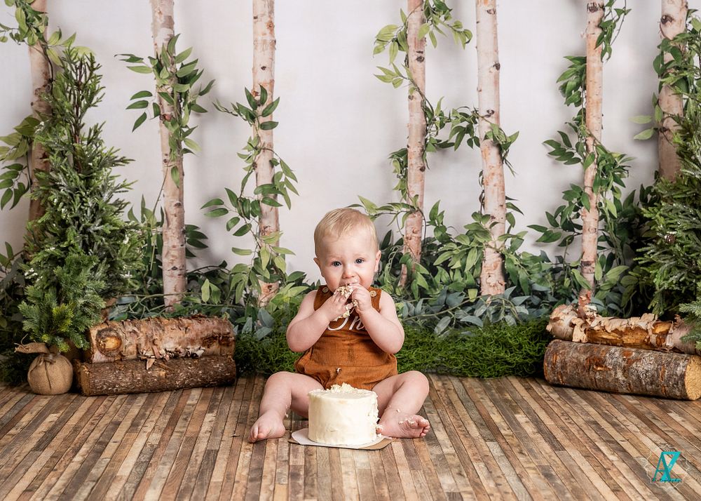 Child in brown romper sits with hands near face against woodland studio backdrop.