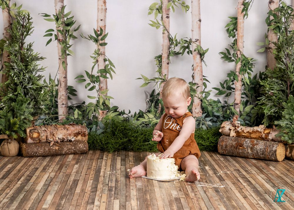 Child in brown overalls plays with white prop box in forest-themed studio setting.