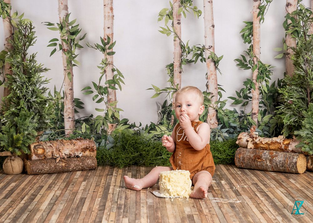 Child in brown overalls sits beside white prop in woodland-themed photo setting.
