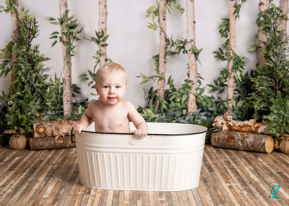 A small child sits in a white metal tub against a rustic backdrop with birch logs and greenery.