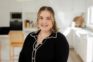 Smiling woman in black blouse poses in modern white kitchen for professional headshot photo.