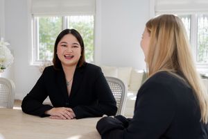 Two professional women in black blazers having a friendly business meeting at a white table in a bright office.
