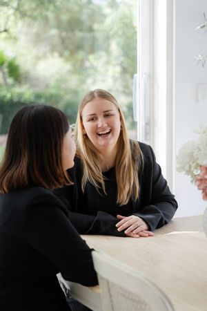 Two young women in black tops laughing and talking together at a table near a bright window.