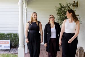 Three women in black outfits walking together on a porch near a for lease sign.