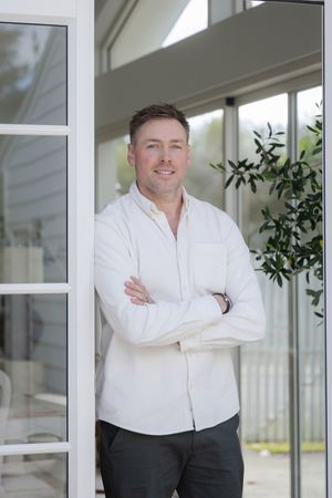 Man in white shirt standing with arms crossed by modern home entrance with glass doors and green foliage.