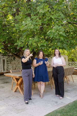Three women smiling outdoors on a patio under a large green tree, dressed in casual elegant outfits.