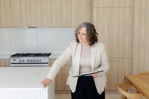 Person gesturing while holding tablet in modern kitchen with light wood cabinets and stainless appliances.