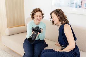 Two people sit on a light colored couch looking at photographs on a digital camera.