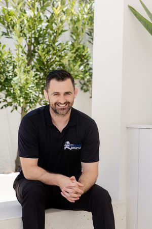 Professional wearing black polo shirt sits on white steps with green foliage in background.