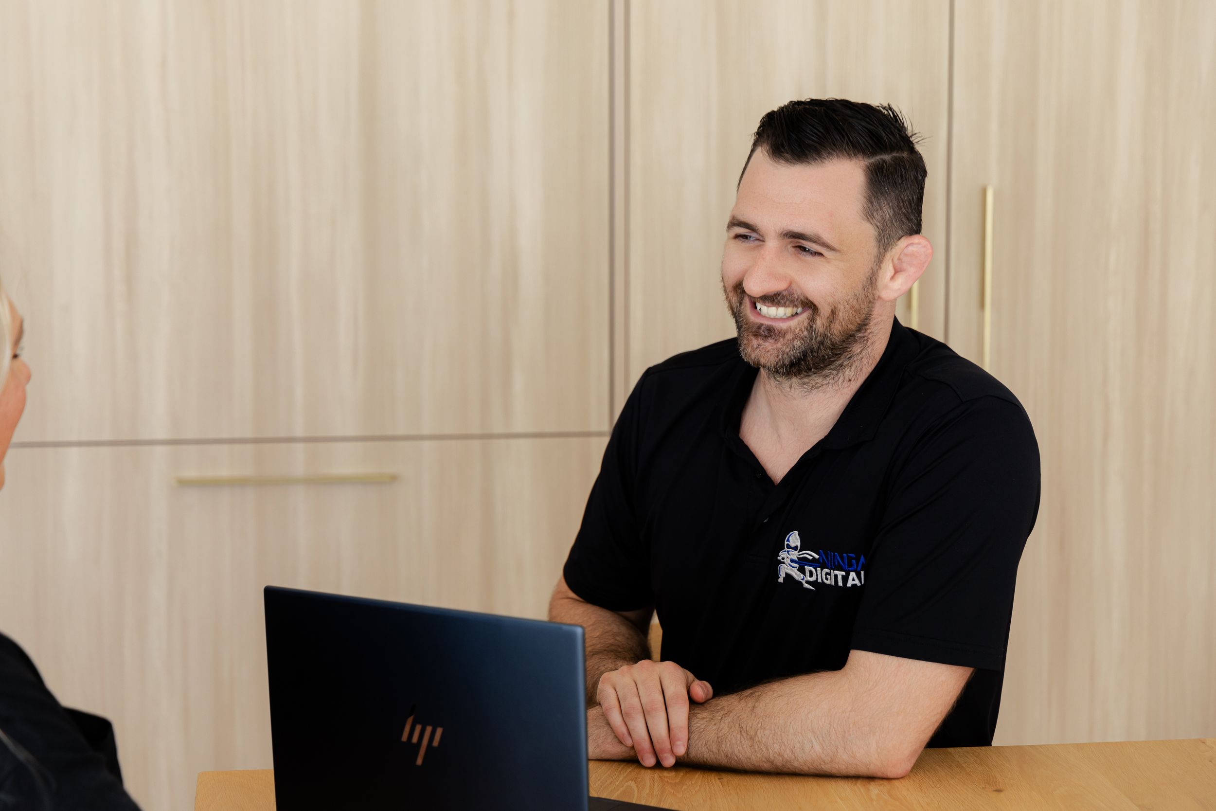 Professional in black polo smiles while working on HP laptop in office setting.