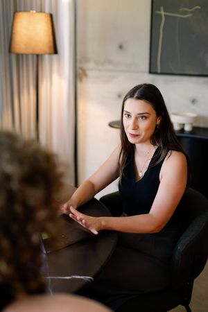 Young woman in black dress sits at dark table in modern interior, gesturing during conversation.