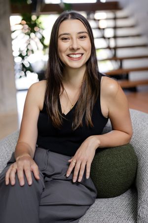Smiling young woman with dark hair sitting relaxed in a modern chair, wearing a black tank top and gray pants.