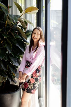 Smiling woman in pink blouse and floral skirt standing by large window next to a rubber plant indoors.