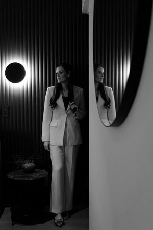 Black and white photo of a woman in a white blazer suit standing near a round mirror with circular wall light.