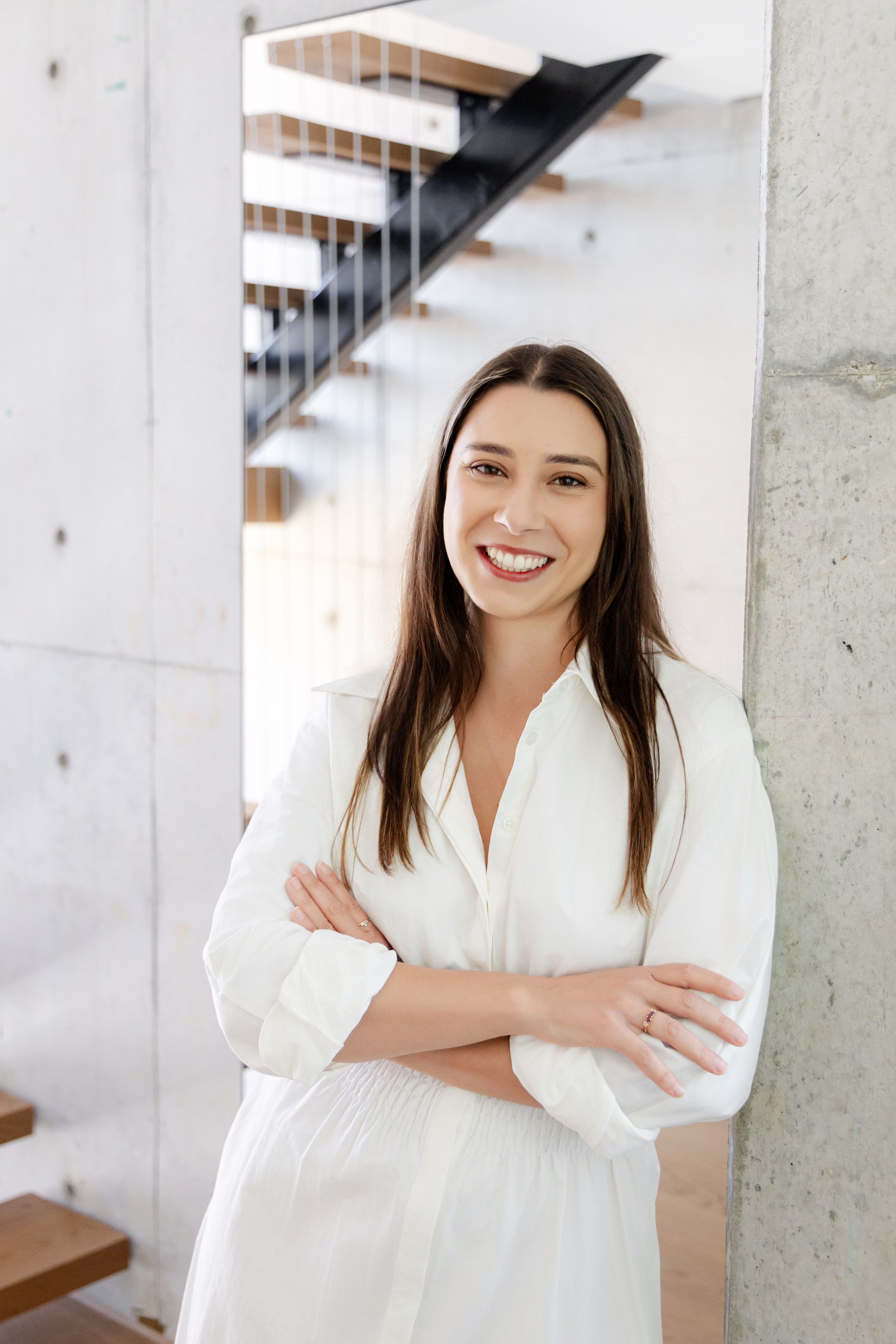 Smiling woman in white dress with arms crossed leaning against concrete pillar near modern staircase.