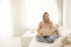 Woman in white room sitting on couch with laptop working while drinking coffee.