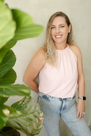 A person in a pink halter top and light jeans stands casually against a white wall with green plant leaves in the foreground.