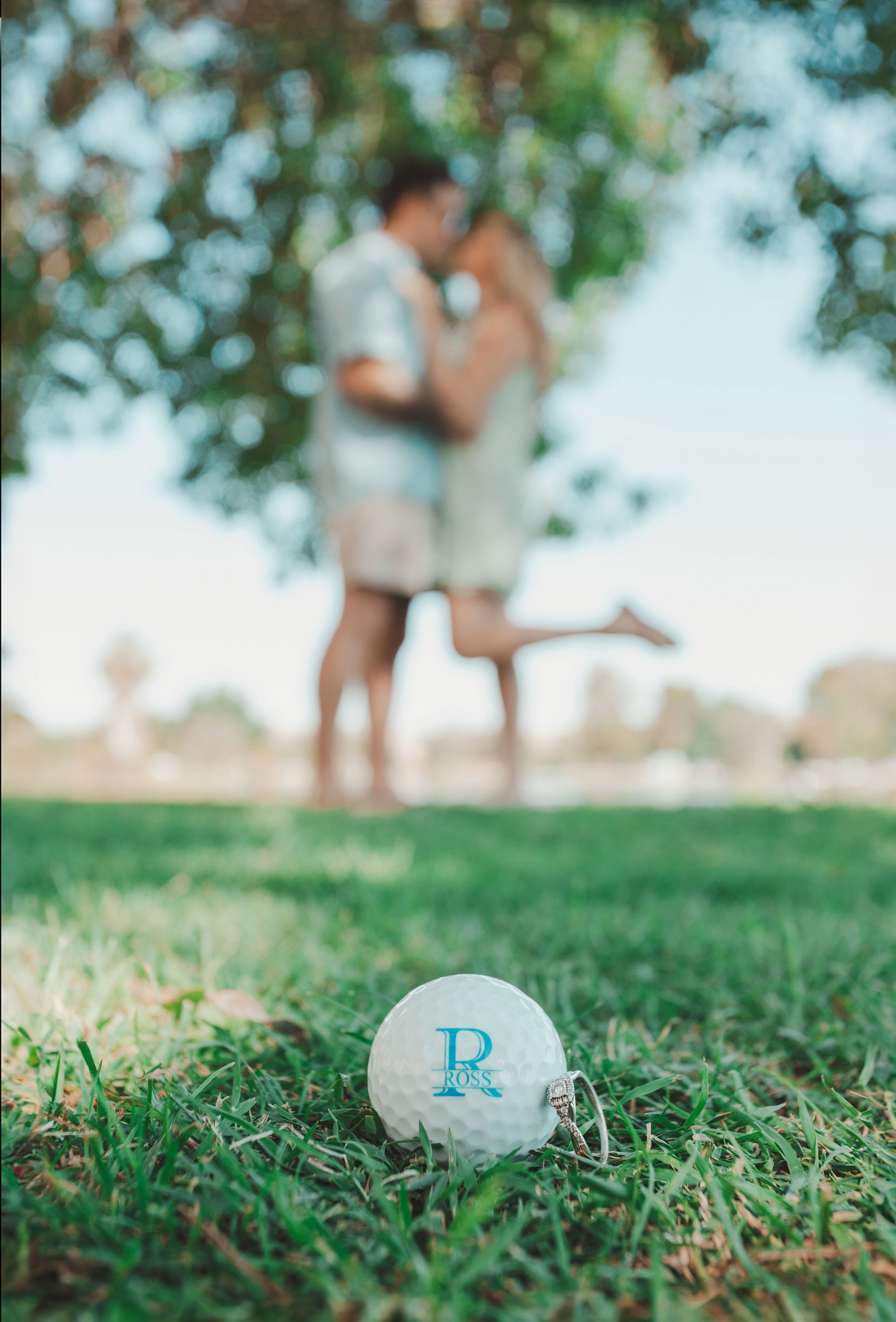 A golf ball with the letter R sits in grass with a blurred couple in the background.