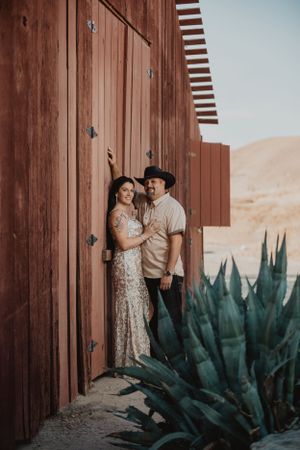 Couple poses against rustic wooden wall with desert landscape and agave plants in background.