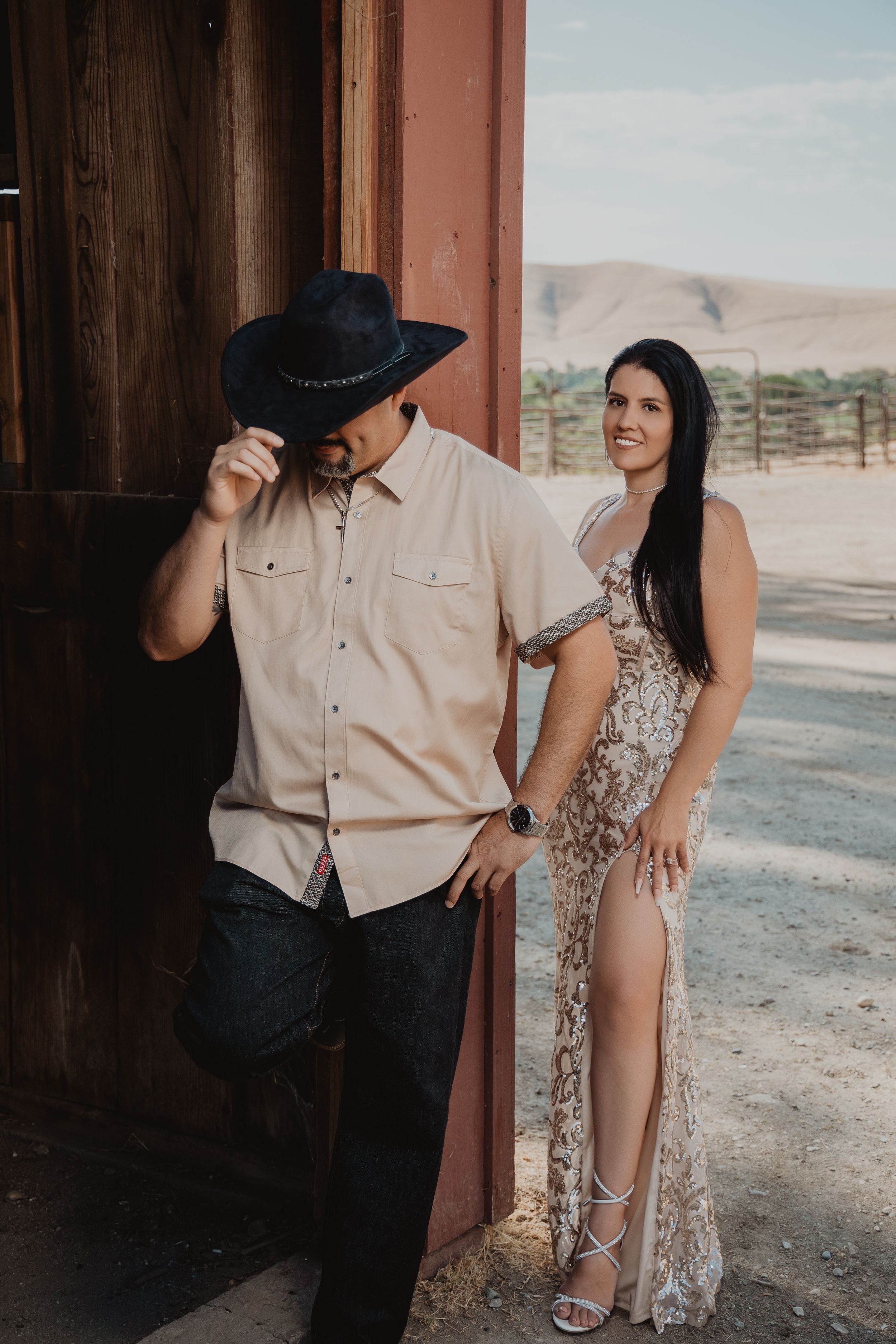 Western-styled couple poses against wooden barn door with desert hills in background.