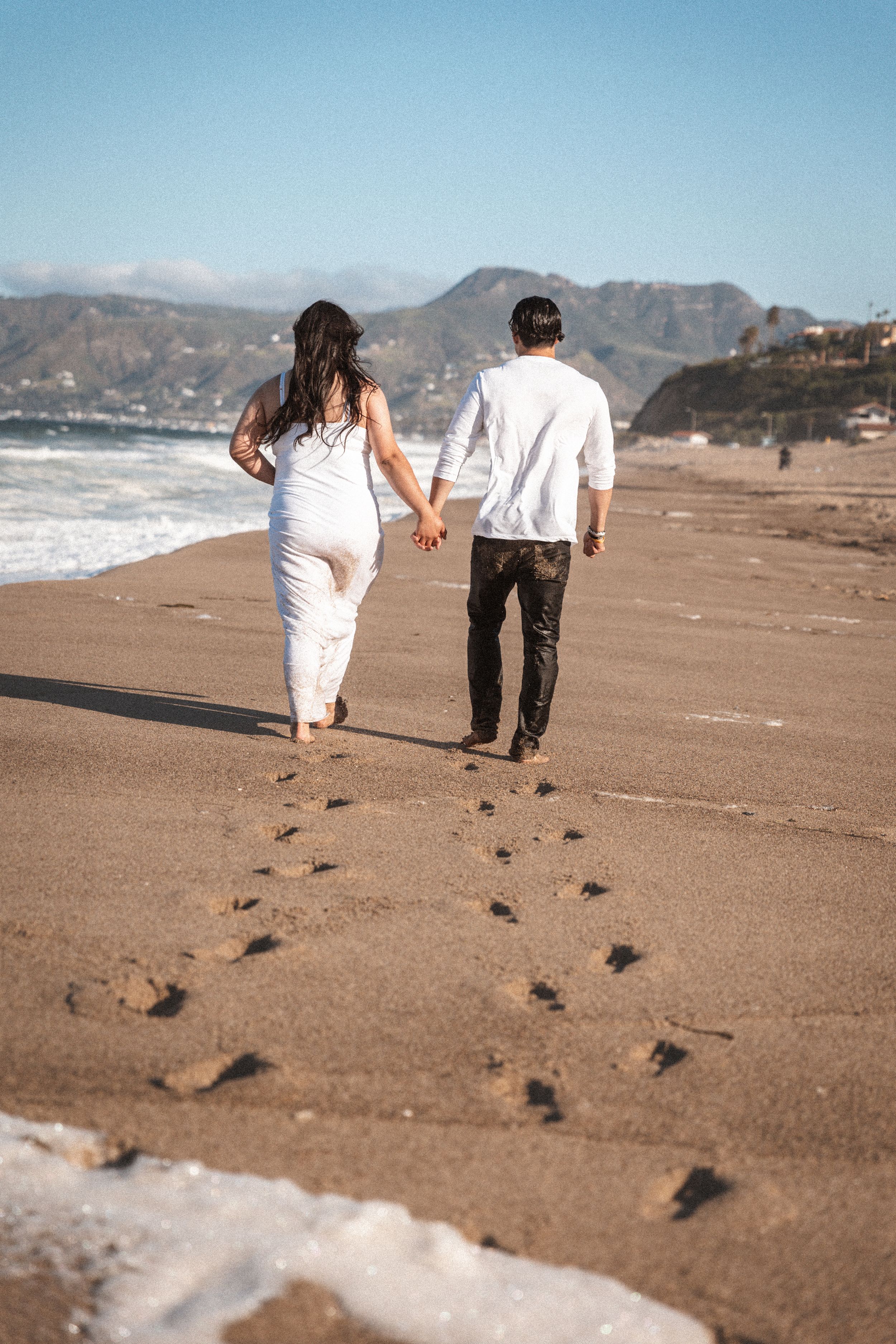 Couple walking along beach leaving footprints in sand with mountains and waves behind them.
