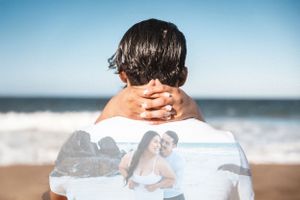 Person holding photo against ocean backdrop with engagement ring visible.