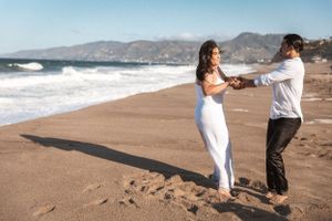 Two people dance together on a sandy beach with mountains and ocean waves in the background.
