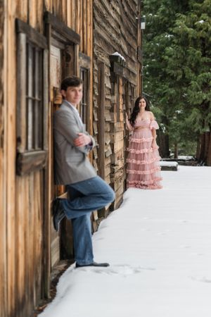 A winter engagement photo featuring a couple by a rustic cabin in the snow.