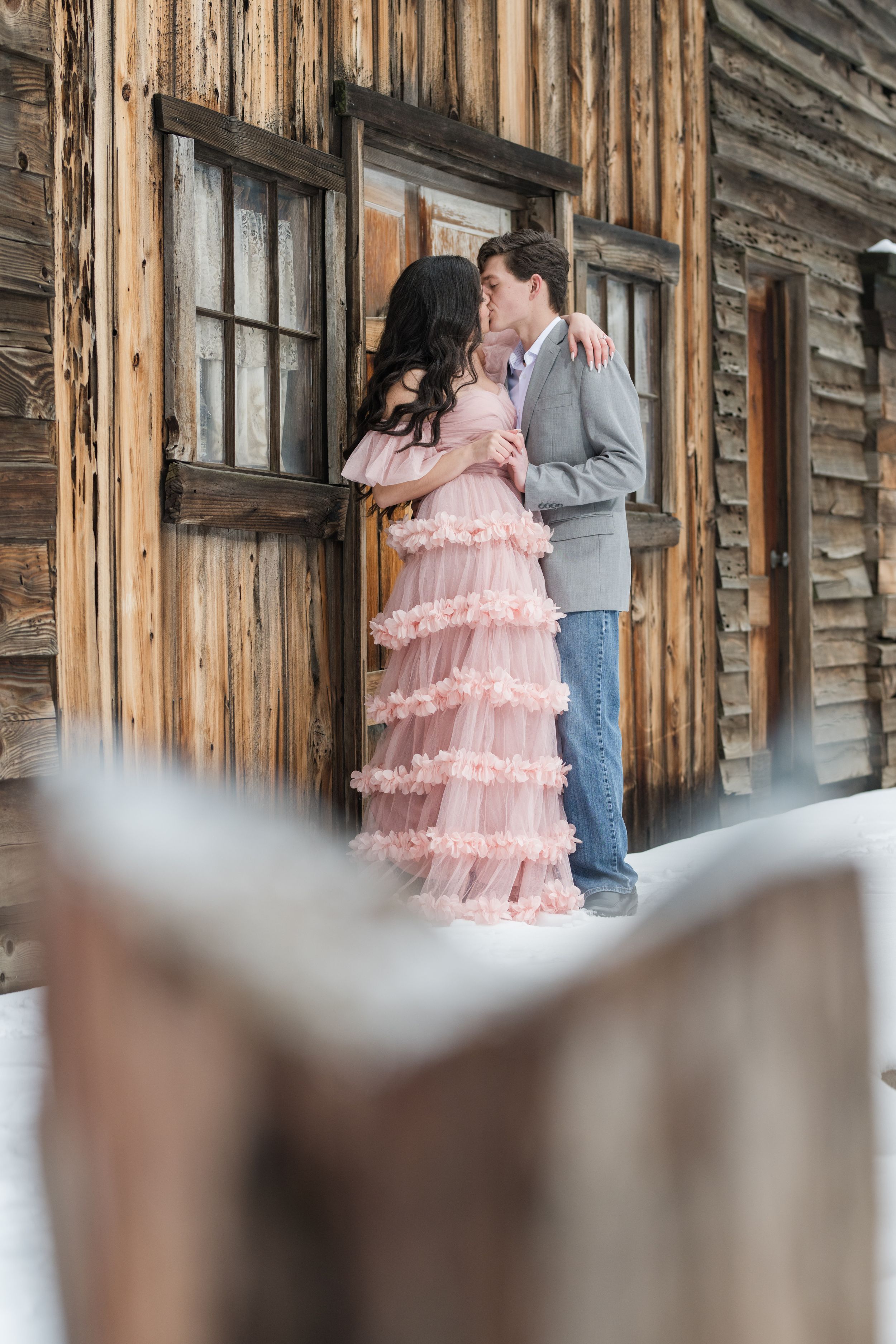 A couple embraces against a weathered wooden building during their winter photoshoot.