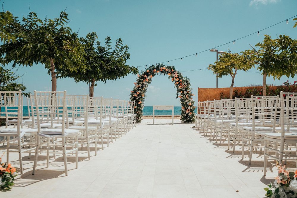 Beachfront wedding setup with floral arch, white chairs, and ocean view under blue skies.