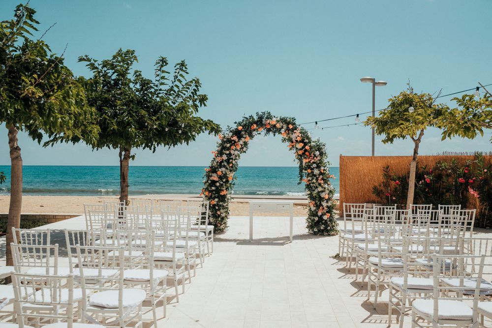 Empty wedding ceremony setup on beach with floral arch and white chairs facing ocean view.