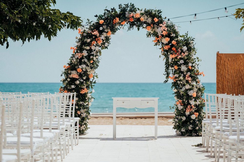 Floral wedding arch with peach and white roses against turquoise ocean backdrop.