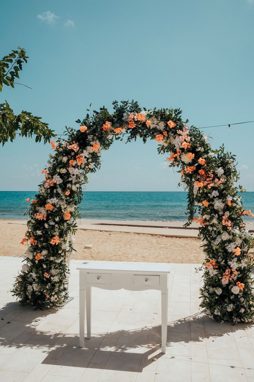 Wedding arch decorated with coral and white flowers on beach with white desk centerpiece.
