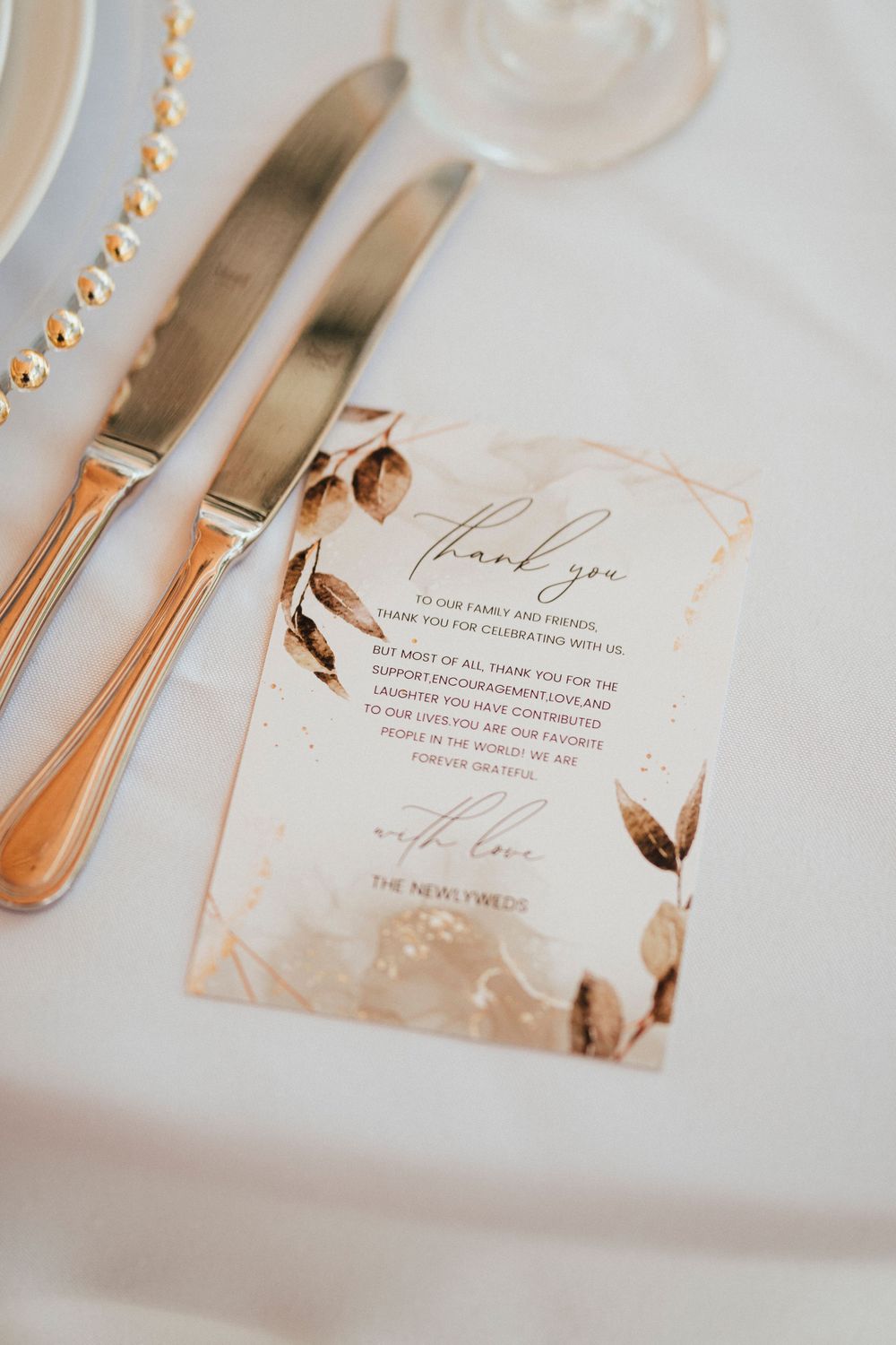 Wedding thank you card with rose gold floral design next to gold flatware on white tablecloth.