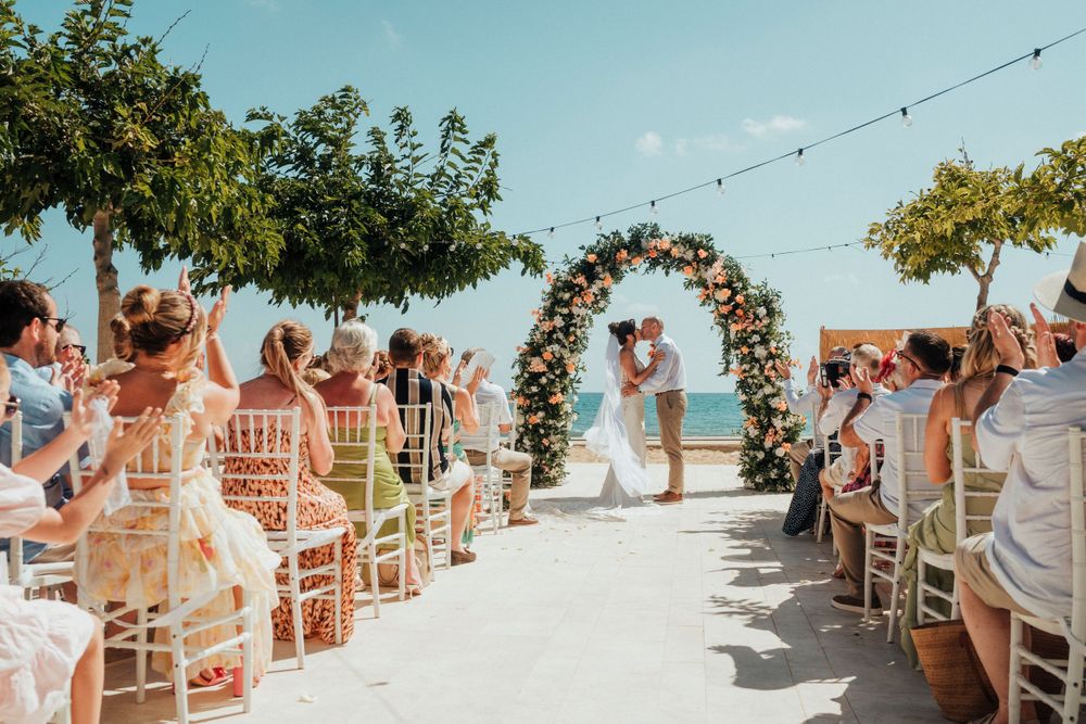 Beach wedding ceremony with floral arch and white chairs as guests watch couple exchange vows.