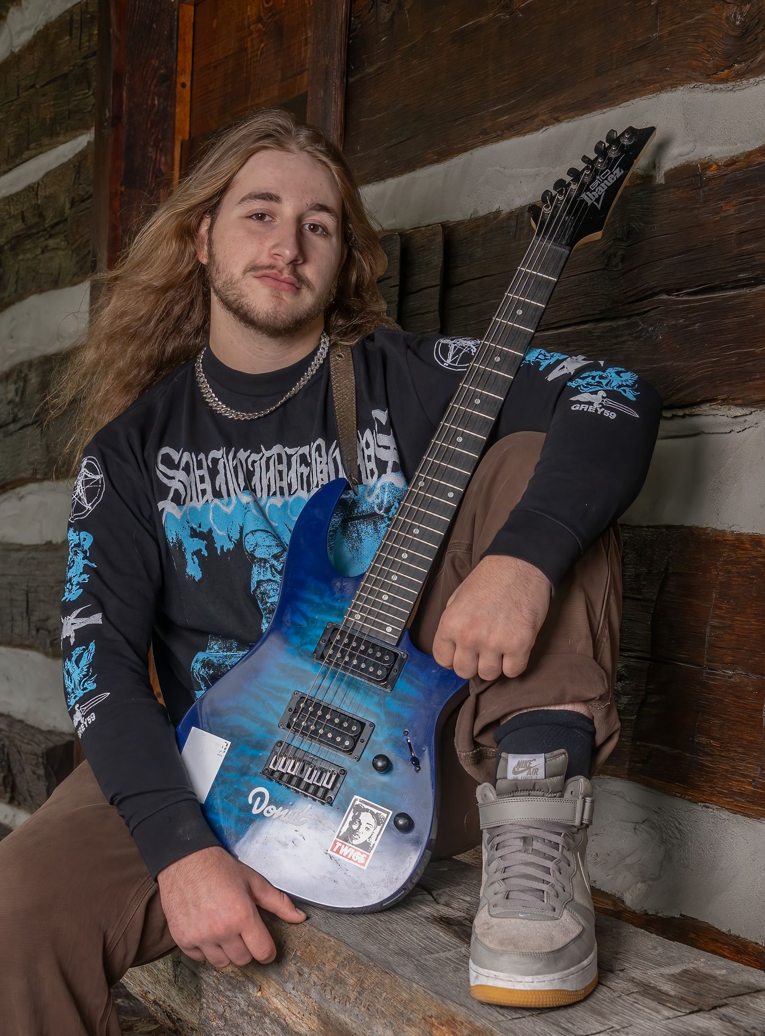 Musician poses with blue electric guitar wearing metal band shirt and brown pants.