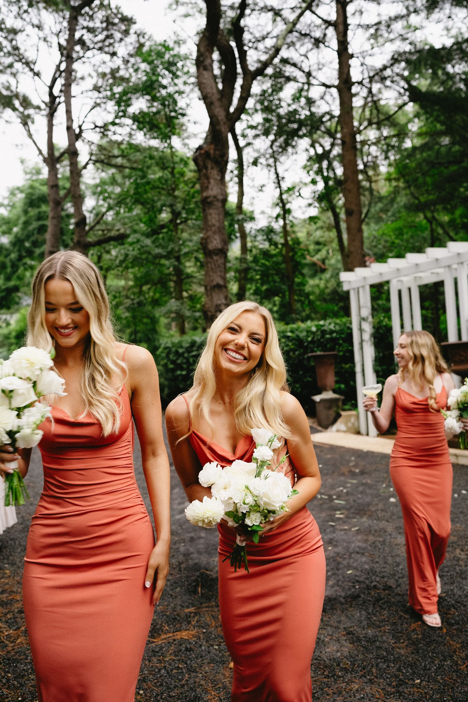 Bridesmaids in rust-colored dresses walking outdoors near white wedding arbor.