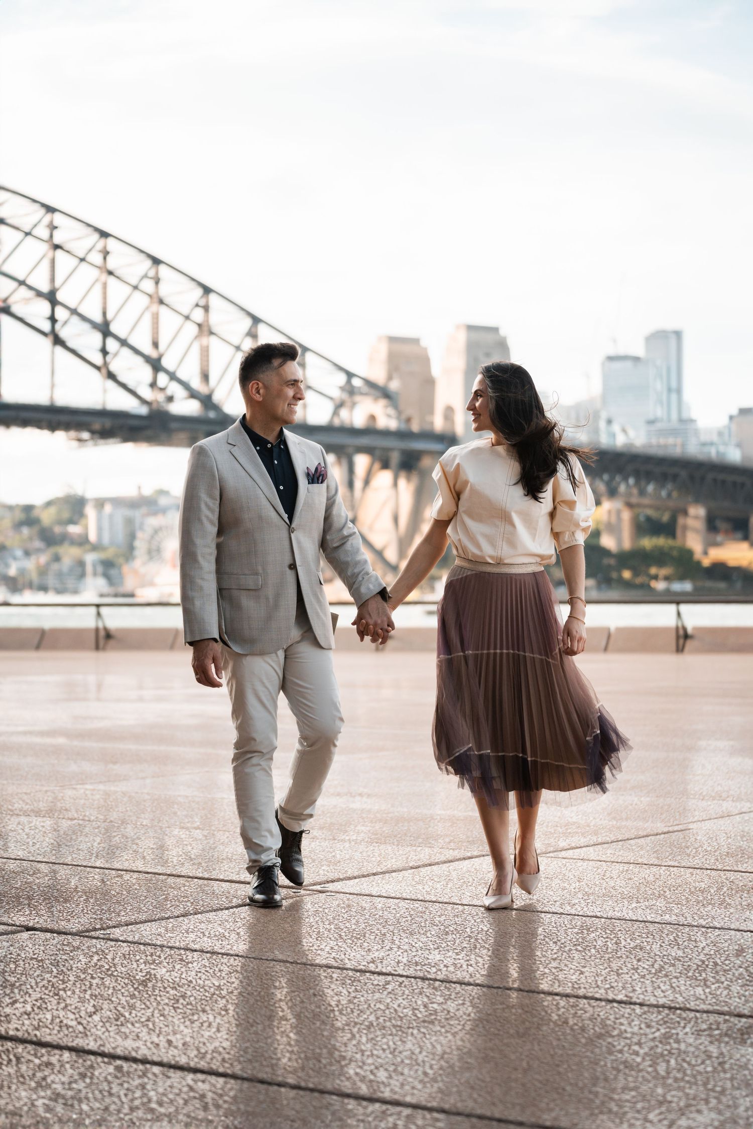 a couple is walking toward the photographer while looking at each other with the Harbour bridge in the background Sydney