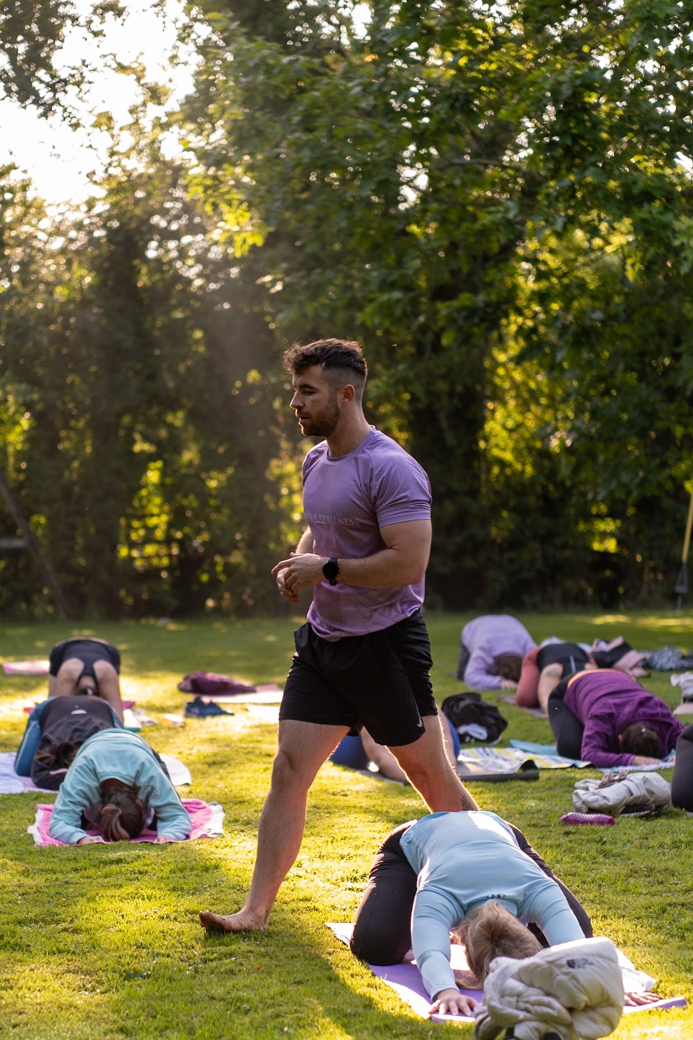 Fitness instructor walking through outdoor yoga class in purple shirt and black shorts.