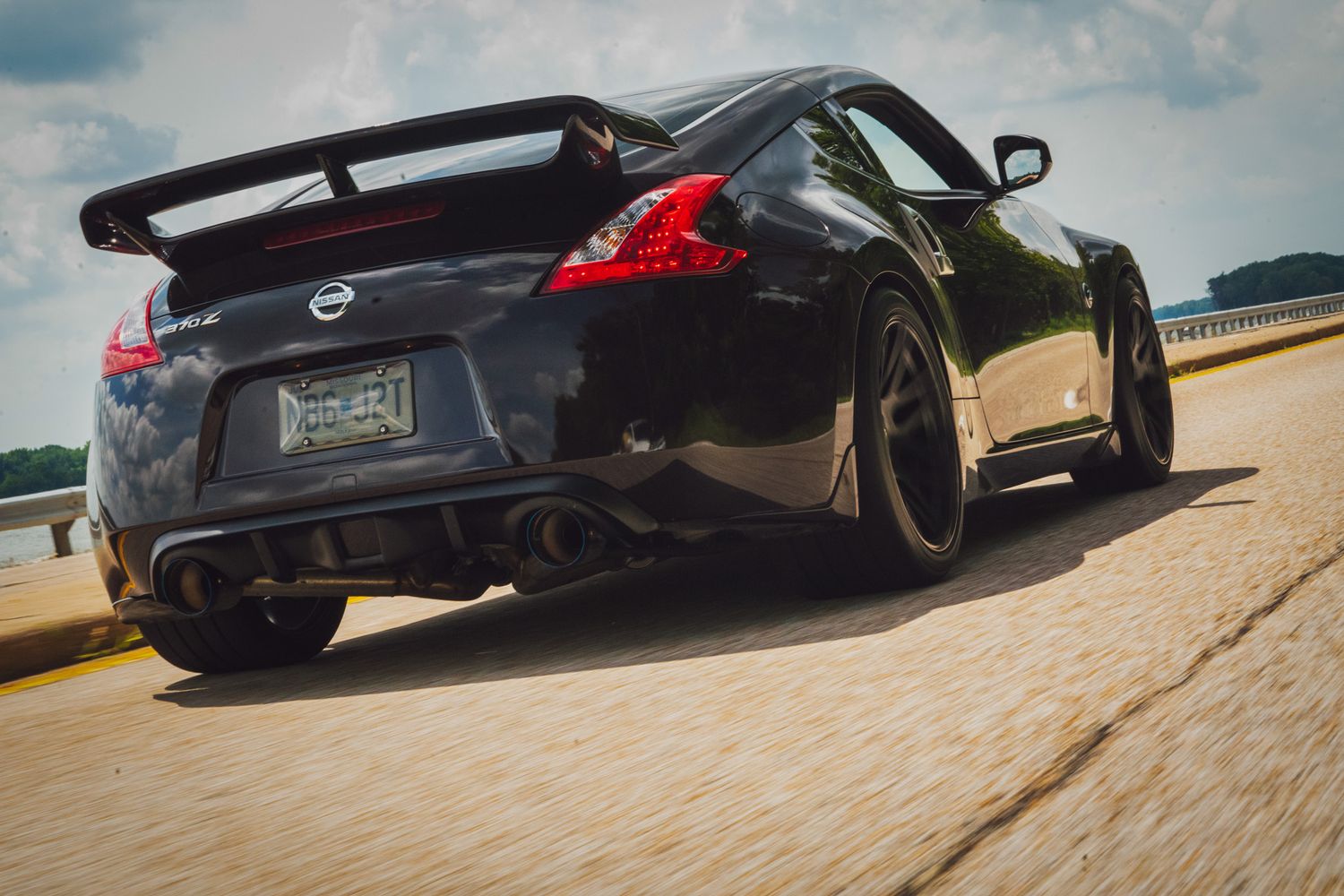 Black Nissan 370Z with custom wide body kit and large rear wing shown from behind on a coastal road.