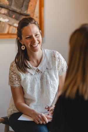 Woman in white lace top smiles warmly during a conversation in an indoor setting.